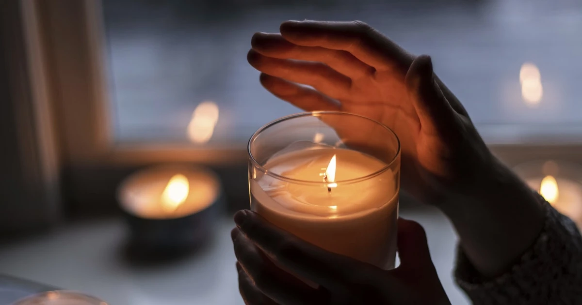 Hands cradling a lit candle in warm, dim light beside a window.
