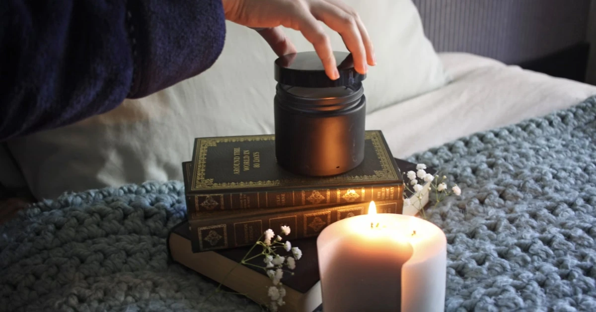 Hand placing a dark cylindrical object on a stack of vintage books beside a lit candle and small flowers.