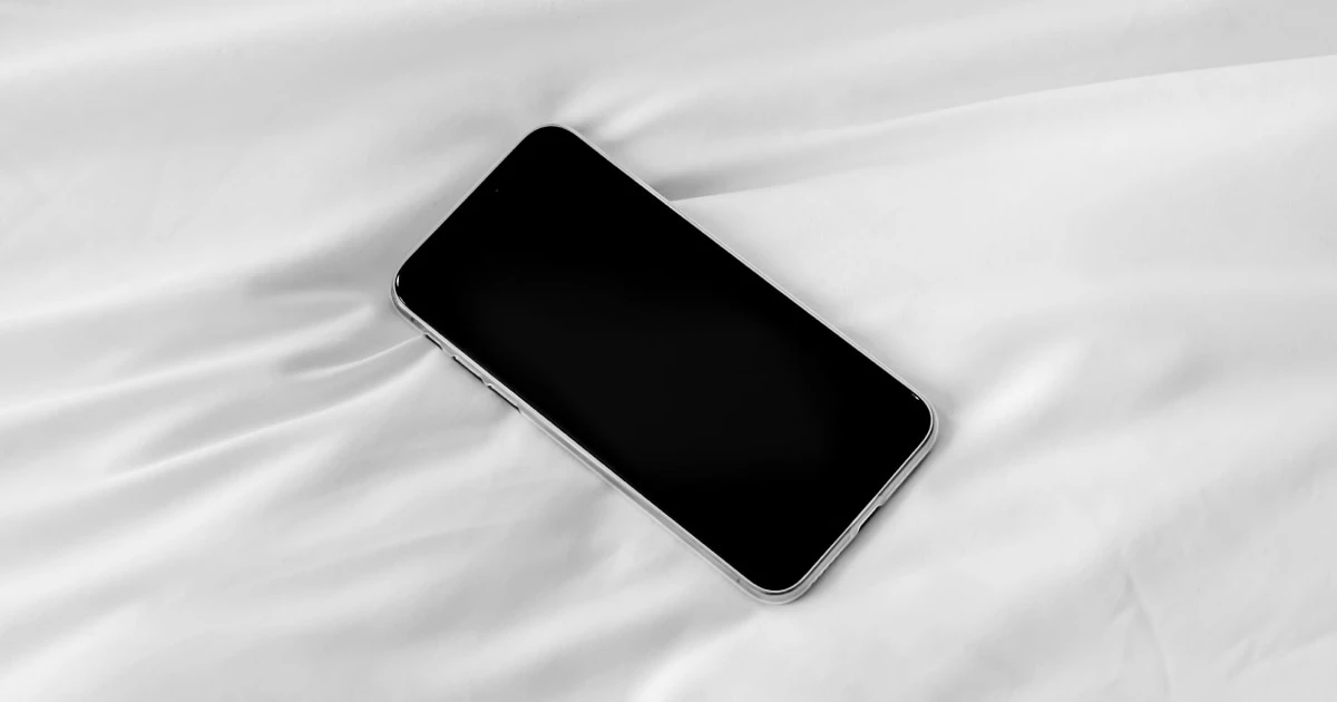 iPhone lying on white bedding with a blue plaid blanket, its lock screen lit.