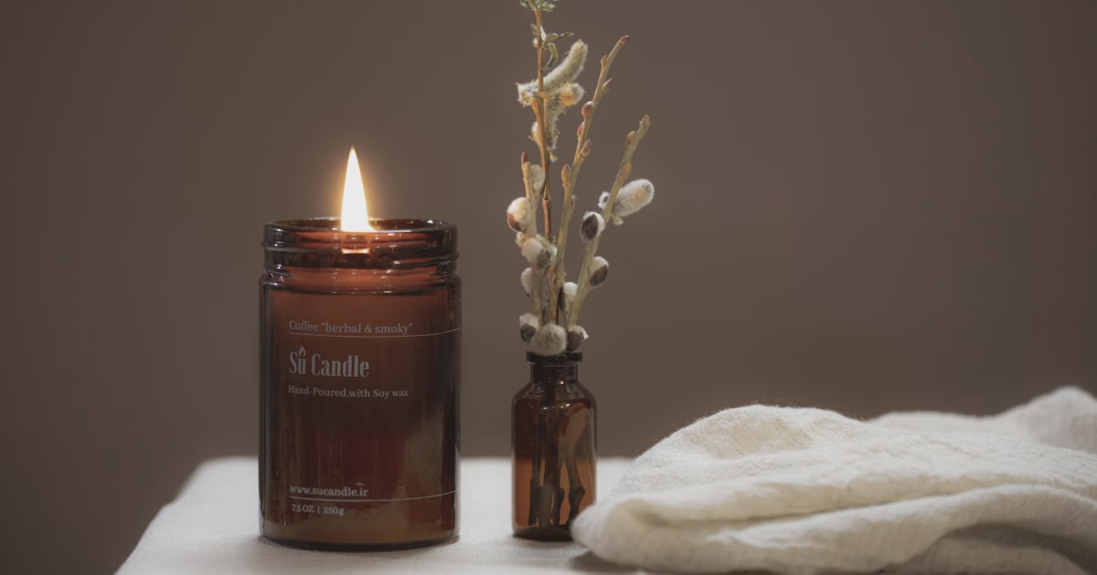 Lit amber candle in a glass jar next to dried flowers and a folded cream cloth on a table.