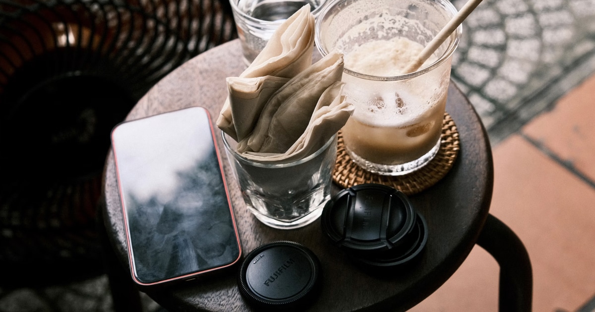 Smartphone on a small round table next to an iced coffee, a folded napkin and a lens cap.