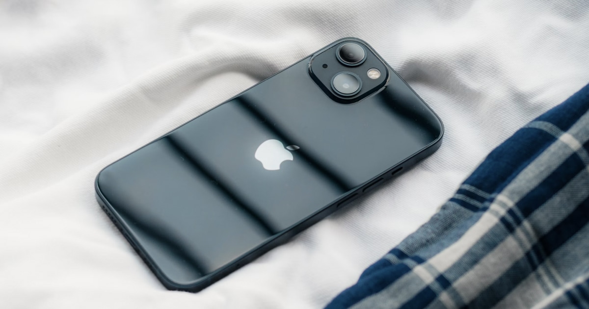 Dark teal iPhone lying face down on white bedding with a blue plaid blanket.